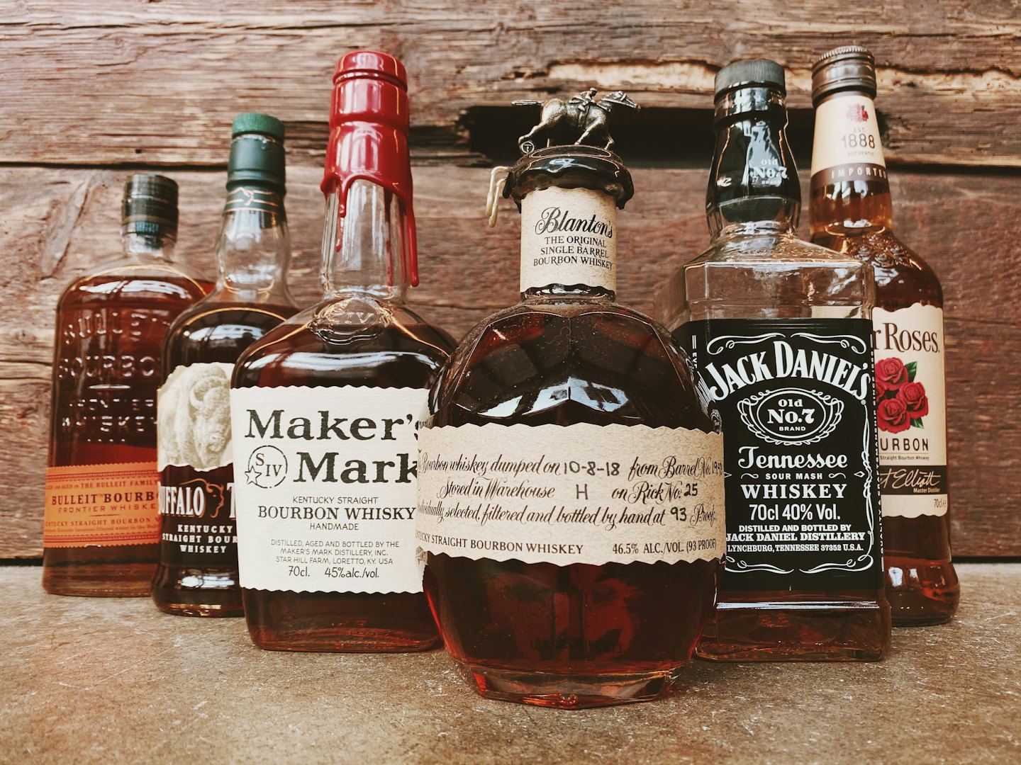 Six bottles of bourbon and whiskey lined up on concrete.